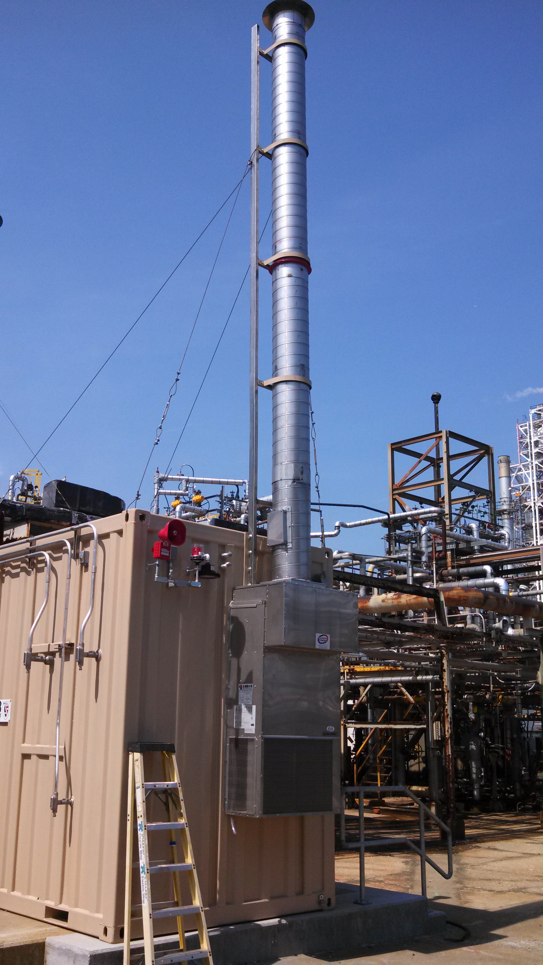 Pressurized equipment shelter at a petrochemical facility, showing galvanized snorkel duct rising approximately 20 feet above the container, wall-mount AC unit, and red pressurization loss alarm horn